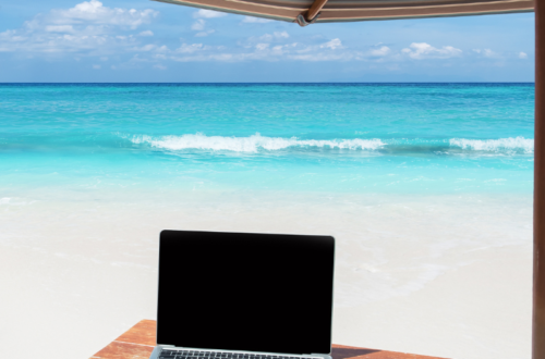 beach waves in the background with a desk and computer under an umbrella in the foreground