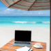 beach waves in the background with a desk and computer under an umbrella in the foreground
