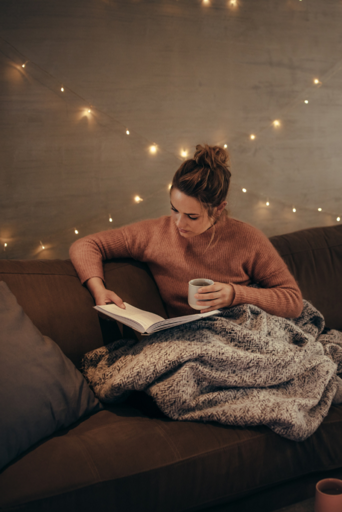 woman reading on a sofa holding a book and a cup of coffee with a blanket over her lap and twinkling lights strung overhead
